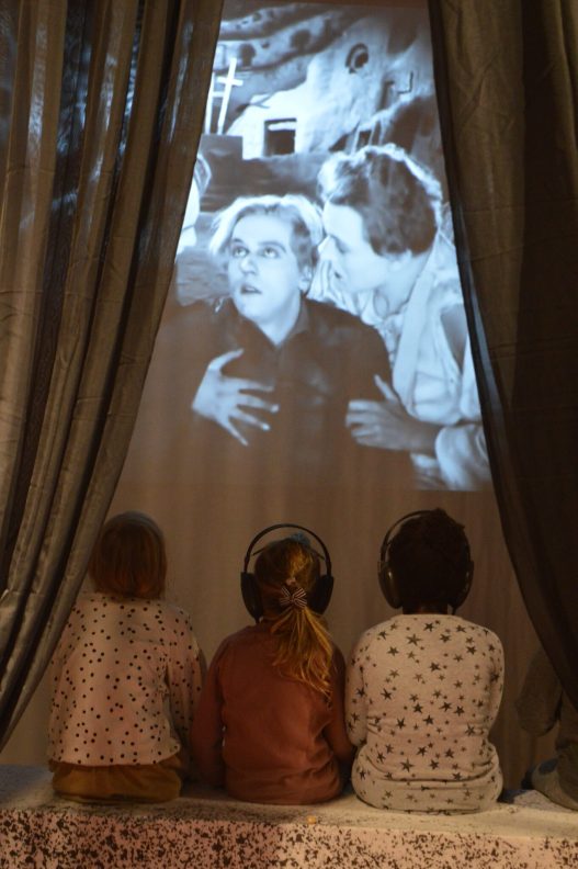 Drei kleine Mädchen von hinten mit Kopfhörern schauen einen alten schwarz-weiß Film, der vertont wurde.