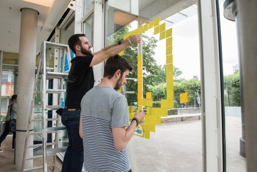 Zwei Männer bekleben das Fenster des Museums mit Post-its und formen daraus Emojis.