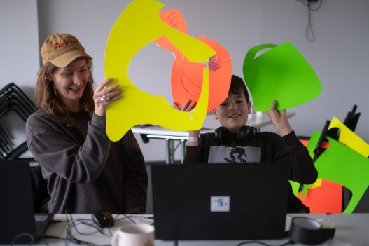 Künstlerin Anna-Luise Lorenz und ein Teilnehmer halten lachend bunte Formen aus Pappe in eine Laptopkamera.