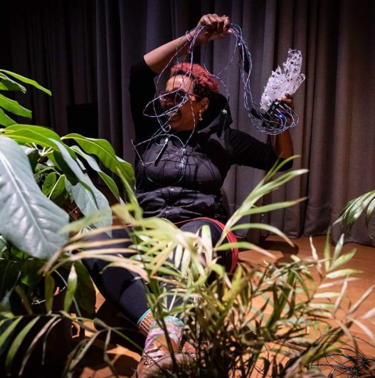Künstler*in Sarah Fartuun Heinze sitzt hinter vielen Pflanzen auf dem Boden und hantiert lachend mit bunten Kabeln.